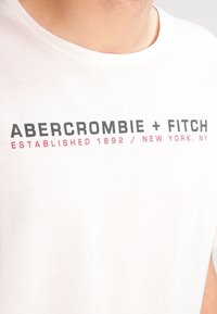 Camiseta blanca de manga corta con texto negro y rosa que dice "ABERCROMBIE + FITCH ESTABLECIDO 1892 / NUEVA YORK, NY." Tejido de algodón suave.