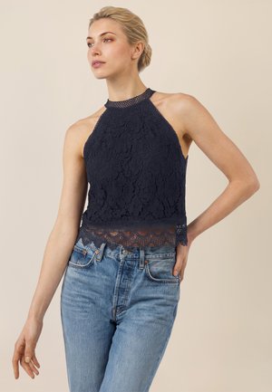 Frau mit blondem Haar, die ein marineblaues Spitzen-Halfter-Top und hellblaue Jeans mit hoher Taille vor einem beigen Hintergrund trägt.