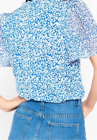 Kortärmad blus i blått med vita abstrakta mönster, tillverkad i ett lätt tyg och med puffärmar, tillsammans med denimbyxor.