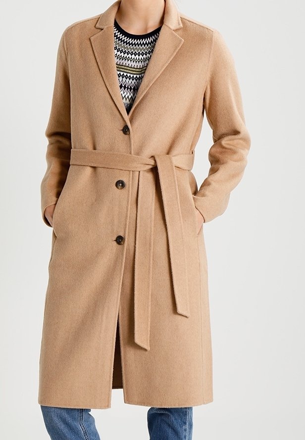 Manteau long en laine beige avec col à revers, fermeture par boutons et ceinture à la taille, porté sur un pull à motifs et un jean bleu.