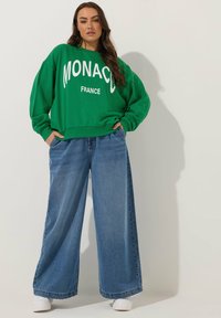Zielona bluza z napisem "MONACO FRANCE" na białym tle, połączona z szerokimi niebieskimi dżinsami i białymi trampkami. Luźny krój i casualowy styl.