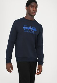 Jūras spēku sporta krekls ar garām piedurknēm, priekšā ar zilu grafisko logo "EA7 Emporio Armani". Mīksts materiāls un brīvs piegriezums.