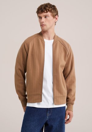 Homme portant une veste zippée marron clair sur une chemise blanche et un jean bleu, face légèrement orientée vers la gauche sur un fond beige uni.