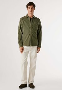 Camisa de color verde olivo, con botones y un bolsillo en el pecho; combinada con pantalones blancos y zapatos marrones. Textura suave, diseño con cuello, ajuste relajado.