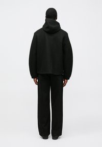 Sort hættejakke lavet af tekstureret stof, med en afslappet pasform og brede ærmer, stylet med løse sorte bukser.