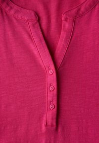 Růžové tričko s krátkým rukávem, které má henley výstřih, čtyři sladěné knoflíky a měkkou, mírně texturovanou látku.