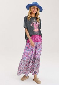 Šedé oversized tričko s růžovou grafikou, spárované s růžovou a multibarevně vzorovanou maxi sukni. Modrý široký klobouk a béžová obuv.