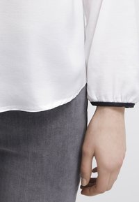 Main reposant à côté d’un pantalon gris, portant un haut blanc à manches longues avec un liseré noir au niveau du poignet, visible de la taille jusqu’au milieu de la cuisse.