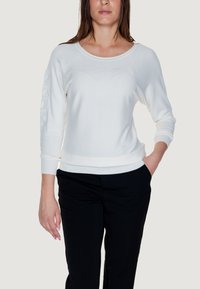 Maglione bianco a maniche lunghe con scollo rotondo e motivo testurizzato sulle maniche; abbinato a pantaloni neri. Tessuto liscio, vestibilità casual.
