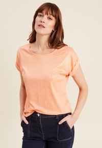 Perzikkleurig top met korte mouwen in een glanzende, zachte stof en een ronde halslijn. Gecombineerd met donkerdenim jeans, met de handen in de zakken.