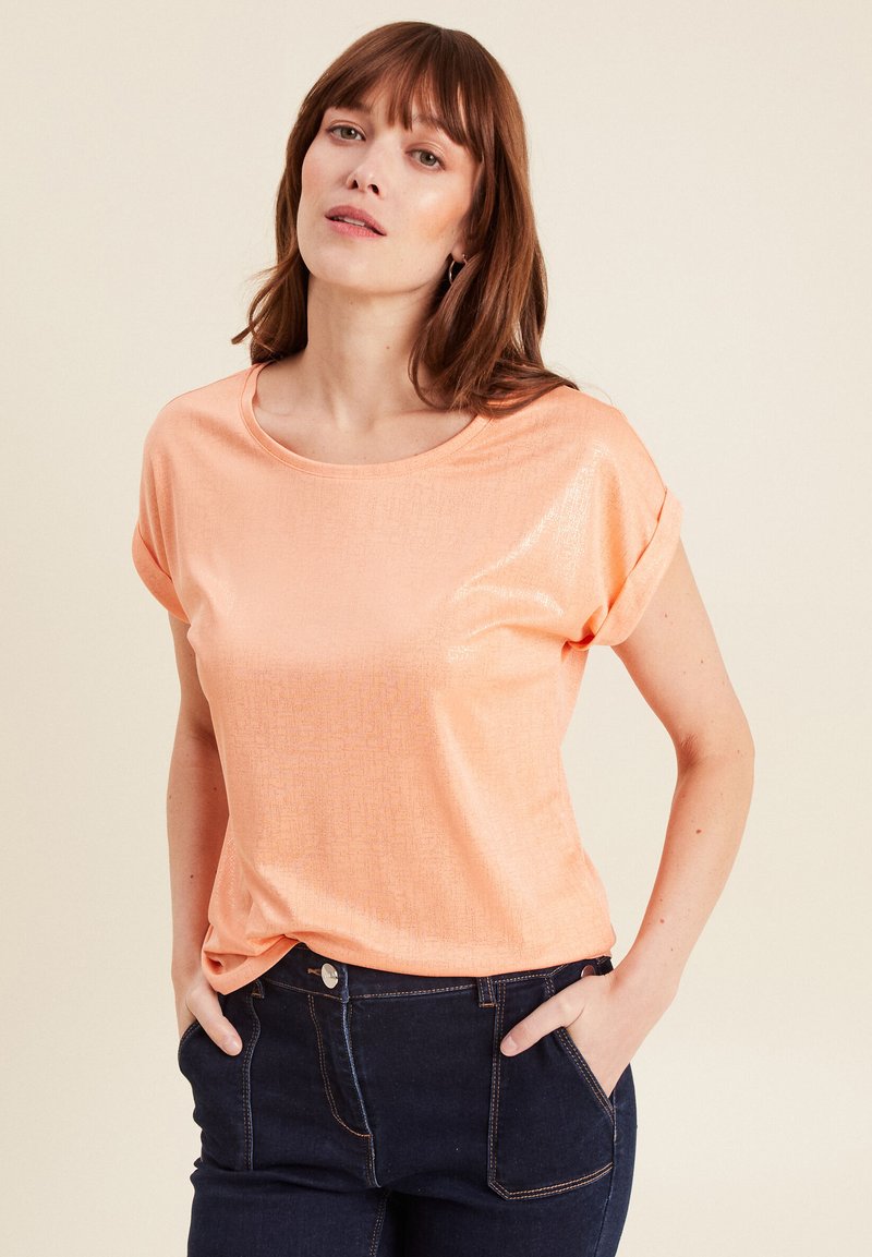 Perzikkleurig top met korte mouwen in een glanzende, zachte stof en een ronde halslijn. Gecombineerd met donkerdenim jeans, met de handen in de zakken.