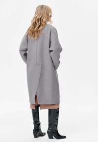 Long manteau en laine gris avec des épaules tombantes, fente dans le dos et poches latérales, porté sur une tenue brune en couches et des bottes noires à talons.