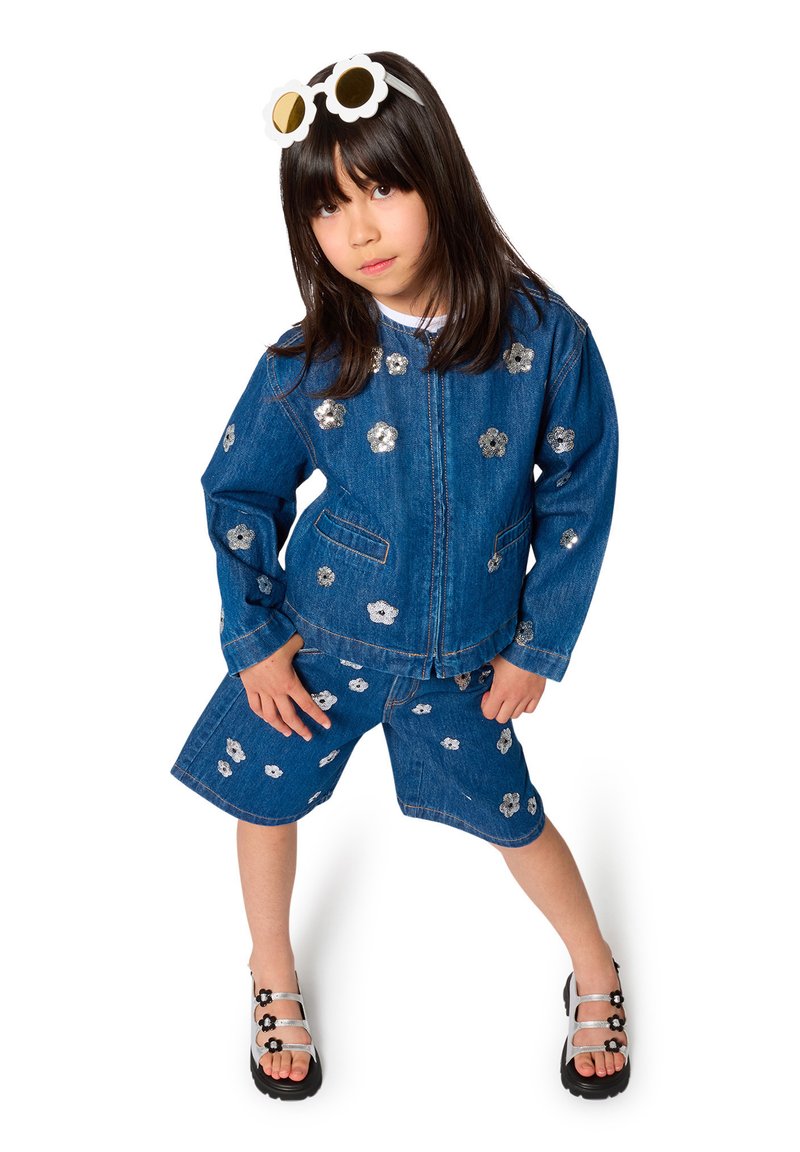 Ensemble veste en jean et shorts ornés de fleurs argentées, accompagné de lunettes de soleil blanches en forme de fleur et de sandales noires.