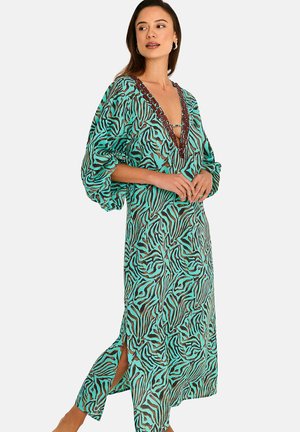 Femme portant une robe imprimée zèbre bleu sarcelle et marron, avec un profond décolleté en V, des manches bouffantes et une fente latérale, regardant vers la gauche.