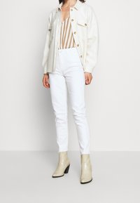 White high-waisted jeans paired with a beige striped bodysuit and an oversized cream button-up shirt; shiny beige ankle boots complete the look.