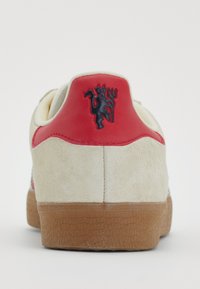 Ténis em vermelho e creme com parte superior em camurça e sola em borracha, apresentando um logo preto no calcanhar e costuras em contraste ao longo das costuras.