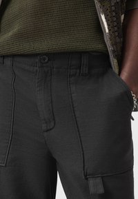 Pantaloni cargo neri con molte tasche, un tessuto strutturato e una vita aderente. Presentano una chiusura con bottone e un aspetto pulito e sartoriale.