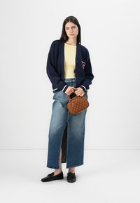 Ναυτικό πουλόβερ με λευκές λεπτομέρειες, κίτρινο μπλουζάκι, ψηλόμεση τζιν φούστα με πλαινές σχισμές, μαύρα παπούτσια και καφέ τσάντα με υφή.