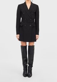 Kobieta w czarnej sukience żakietowej zapinanej na podwójne guziki z paskiem, w połączeniu z czarnymi, skórzanymi kozakami do kolan na jednolitym tle.