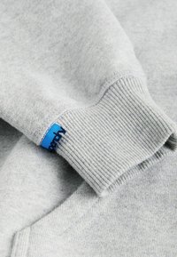 Tejido de sudadera gris con puños de canalé que presentan una etiqueta azul. Superficie texturizada, acabado suave y un diseño clásico redondeado.