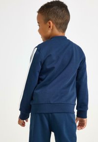 Jongen met kort haar, gekleed in een marineblauwe trainingsoutfit met witte strepen op de mouwen, staand en zijwaarts gericht tegen een witte achtergrond.
