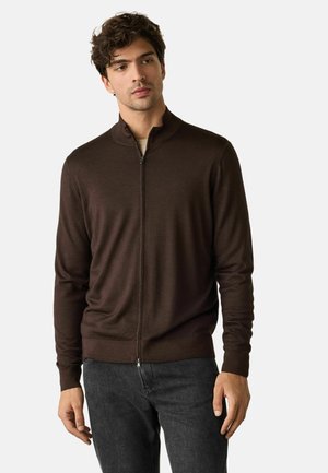 Ung mand iført en mørkebrun lynlåstrøje og mørkegrå jeans står foran en ensfarvet lys baggrund.