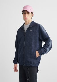 Νεαρός άνδρας που φοράει ένα μπλε σκούρο μπουφάν Puma με φερμουάρ, μαύρο παντελόνι με λεπτομέρειες με καρφιά και ροζ καπέλο baseball Puma, στέκεται με το ένα χέρι στην τσέπη.