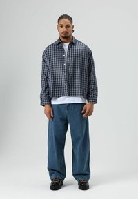 Kariertes Overshirt in Marineblau und Grau mit Kragen, Knopfverschluss und lockerem Schnitt, kombiniert mit hellblauen Jeans mit weitem Bein und schwarzen Schuhen.