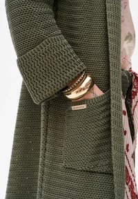 Olijfgroene gebreide jas met een gestructureerd patroon, voorzien van een patchzak en een merketiket. Inclusief een glanzende gouden armband om de pols.