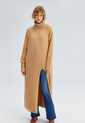 Lange, beige gebreide jurk met hoge kraag, split aan de zijkant en geribde manchetten, gedragen over blauwe flared jeans. Glad textuur en relaxte pasvorm.
