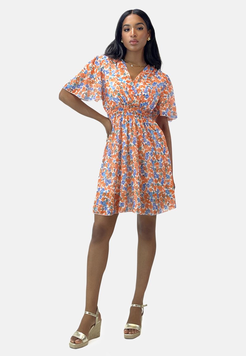 Bloem jurk met een oranje, blauwe en witte print, korte mouwen, een cinched taille en een uitlopende rok. Gecombineerd met gouden sleehakken.