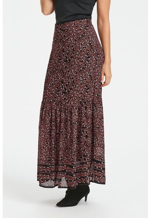 Femme portant une jupe longue noire taille haute avec imprimé floral rose et ourlet à volants, associée à des bottines à talons noires.