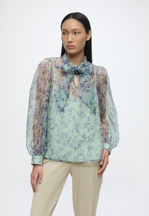 Frau mit glatten schwarzen Haaren, die eine transparente Blumenbluse mit gebundener Schleife am Hals und beige Hose trägt, steht vor einem weißen Hintergrund.