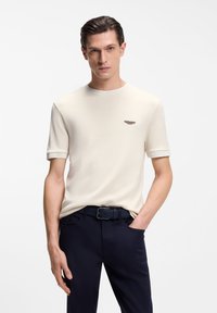 Κρεμ κοντομάνικη μπλούζα με στρογγυλή λαιμόκοψη, με ένα μικρό καφέ λογότυπο στο στήθος και αντίθετες ριμπ μανσέτες. Συνδυάζεται με σκούρο παντελόνι.