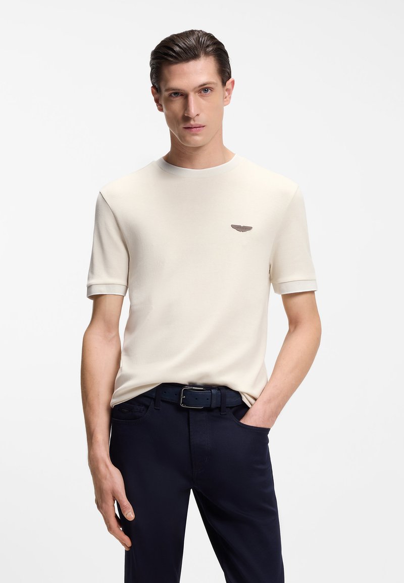 Κρεμ κοντομάνικη μπλούζα με στρογγυλή λαιμόκοψη, με ένα μικρό καφέ λογότυπο στο στήθος και αντίθετες ριμπ μανσέτες. Συνδυάζεται με σκούρο παντελόνι.