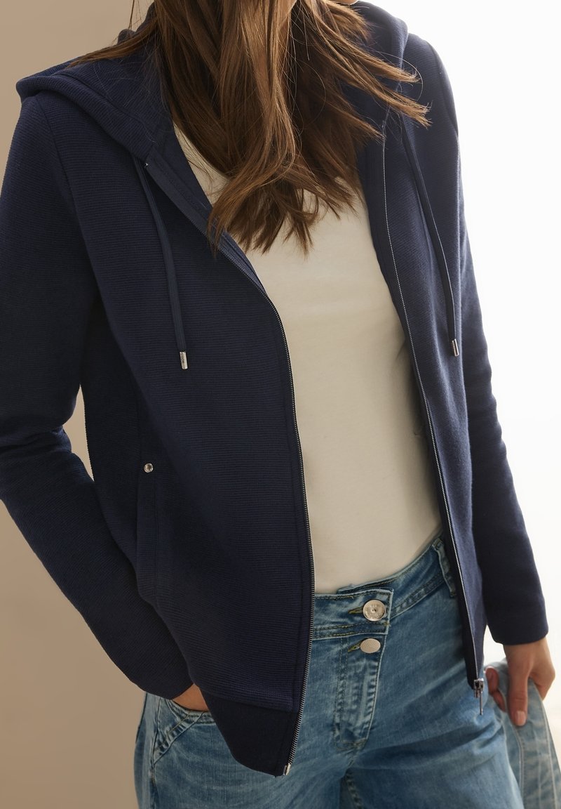 Personne portant un sweat à capuche bleu marine avec fermeture éclair et cordons, par-dessus une chemise blanche et un jean bleu clair, debout avec une main dans la poche.