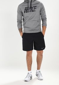 Haut à capuche gris pour homme avec le logo Nike noir, associé à un short noir. Chaussures de sport blanches avec des accents noirs. Texture de tissu lisse.