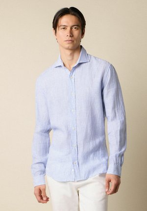 Mann trägt hellblau gestreiftes, langärmliges Hemd mit Knöpfen und weiße Hose, steht vor beigem Hintergrund.