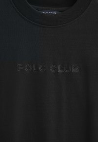 Camiseta de algodón negra con cuello redondo acanalado, que presenta un logotipo tonal "POLO CLUB" bordado en la parte delantera. Tejido con textura y variación de color.