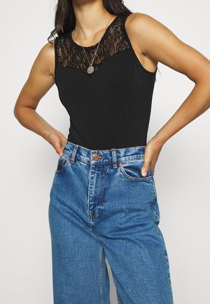 Kobieta w czarnym topie bez rękawów z koronkowym dekoltem i niebieskich dżinsach z wysokim stanem, ręce oparte na biodrach, nosi naszyjnik z okrągłą zawieszką.