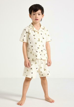 Młody chłopiec stoi boso, ubrany w kremową koszulkę z krótkim rękawem i szorty z małymi wzorami gałązek oliwki.