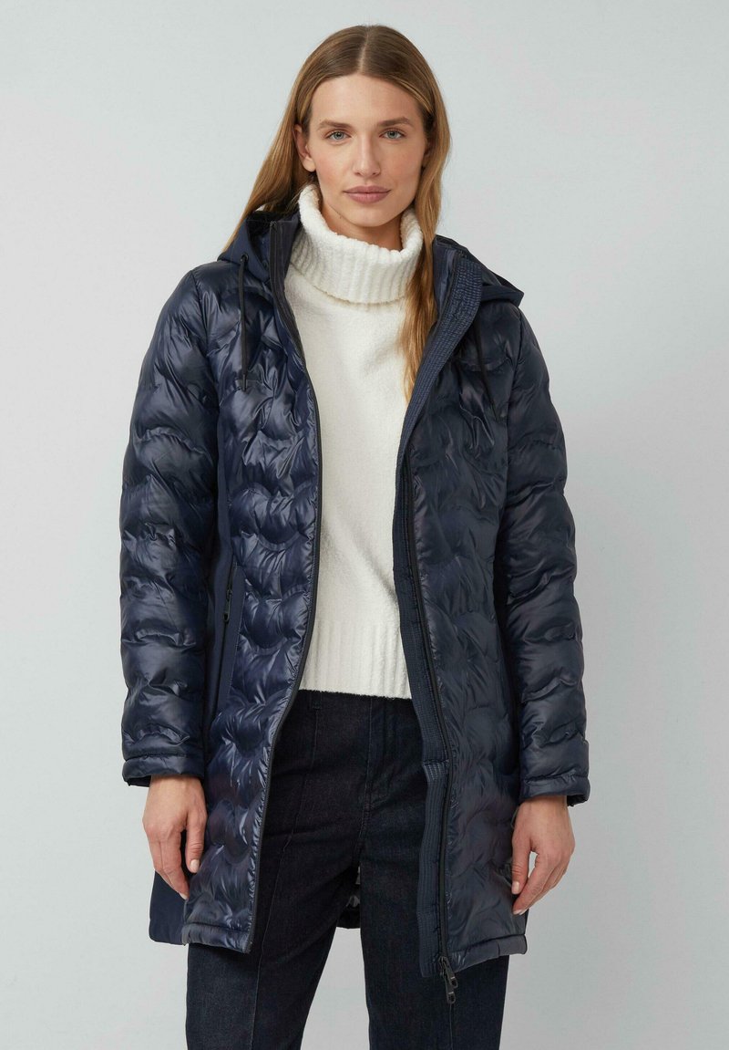 Mornarsko modra puhovka s kvadratnim vzorcem, kapuco in sprednjim zadrgo. Nosi se čez belo rebrasto majico, v kombinaciji s temnimi hlačami. Gladek, sijoč material.