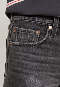 Mörkgrå denimjeans med en slitnitsad yta, kopparnitar och en liten framficka. Sytt detaljer syns längs kanterna.