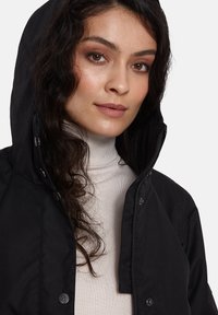 Juodas gobtuvas striukė su užtrauktuku ir spaustuku, pasižyminti lygiu paviršiumi ir šiuolaikišku dizainu. Dėvima virš šviesiai smėlio spalvos golfuko.