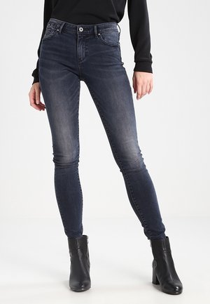 Personne portant un jean skinny bleu foncé et des bottines noires, debout devant un fond blanc uni.
