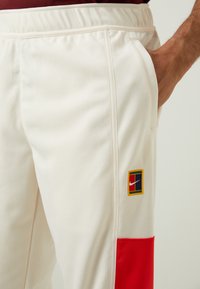Nike joggebukser i kremfarge med rødt fargeblokk. Har elastisk midje, sidelommer og et logo-patch i blått, grønt, gult og rødt.