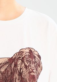 Valkoinen t-paita, jossa edessä yksityiskohtainen ruskea leijonakuva. Kangas näyttää sileältä ja siinä on pyöreä pääntie.