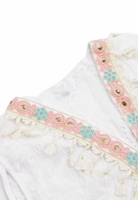 Abito bianco ricamato con motivi floreali colorati in rosa e turchese, con nappine decorative lungo l'orlo e decorazioni.