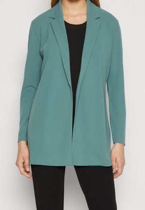 Nainen, jolla on päällään meripihansininen avoin bleiseri mustan topin ja mustien housujen päällä, seisoo yksinkertaisen valkoisen taustan edessä.