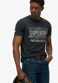 Zwarte katoenen t-shirt met een grote grijze grafische print en logo. Gecombineerd met een blauwe spijkerbroek en een bruine leren riem. Inclusief een geruit overhemd.